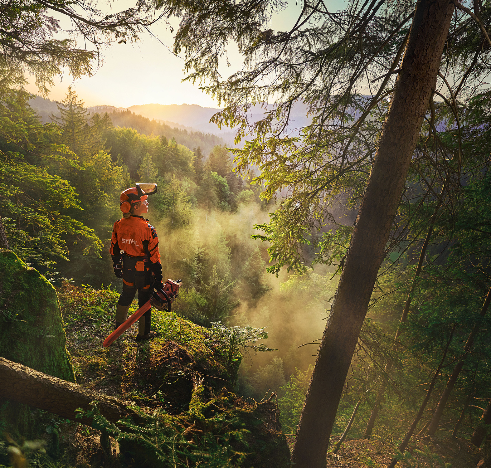 Une femme se tient au bord d'une falaise dans une forêt d'un vert saturé. Elle porte des vêtements de travail STIHL orange, le casque ADVANCE ProCom et tient une tronçonneuse STIHL MS.