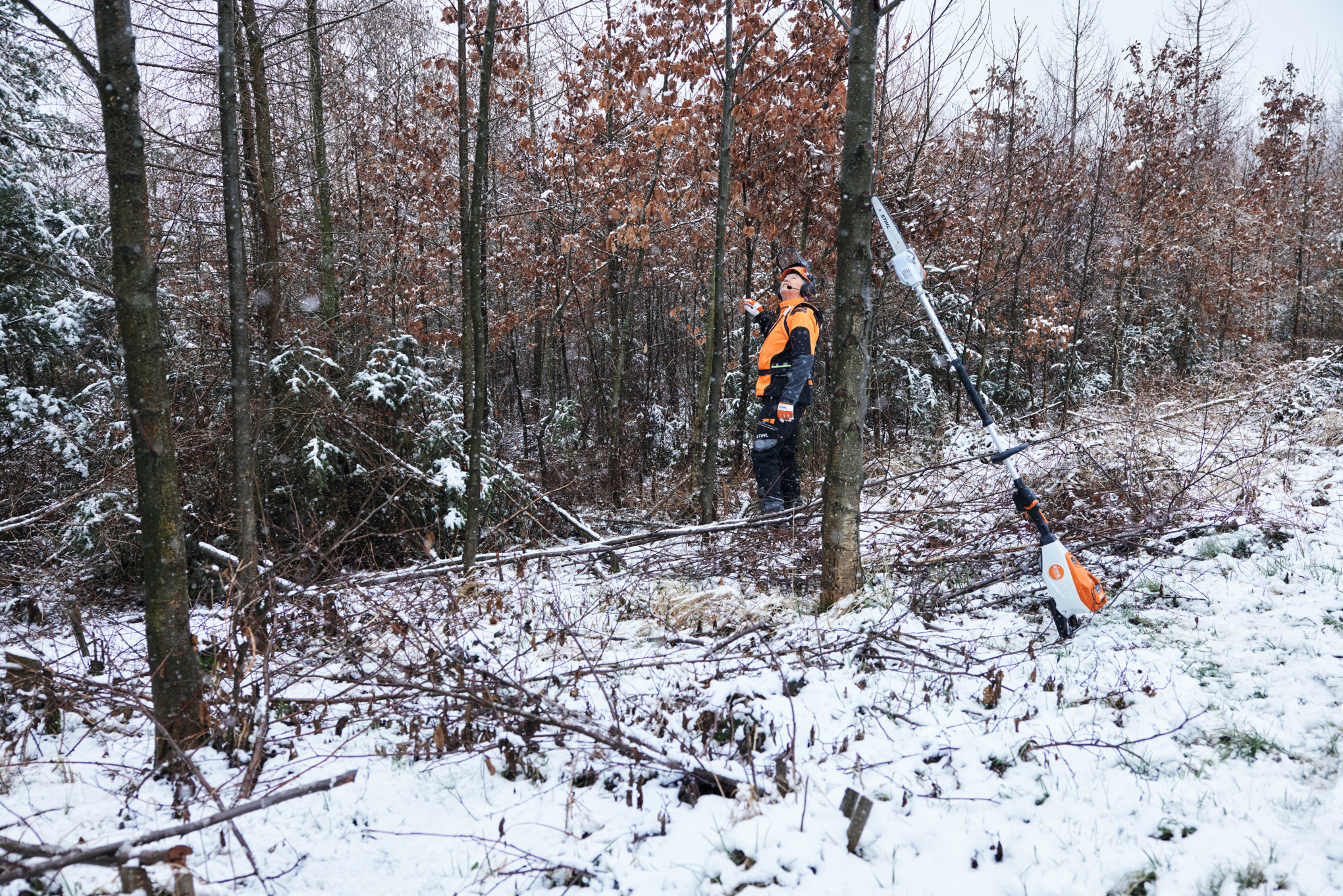 Un homme travaillant en équipement de protection dans une forêt enneigée, au premier plan une perche d’élagage à batterie STIHL HTA 150 posée contre un tronc d’arbre