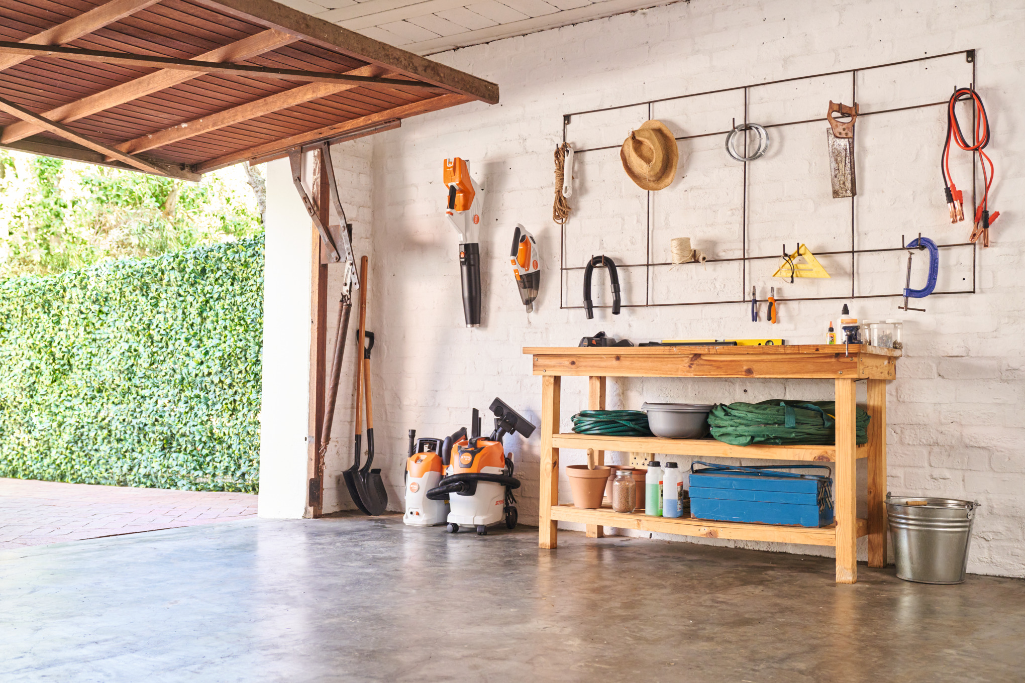 des outils à batteries STIHL sont rangés dans un garage près d'un établi ou accroché au mur, la porte du garage est ouverte et donne sur un jardin