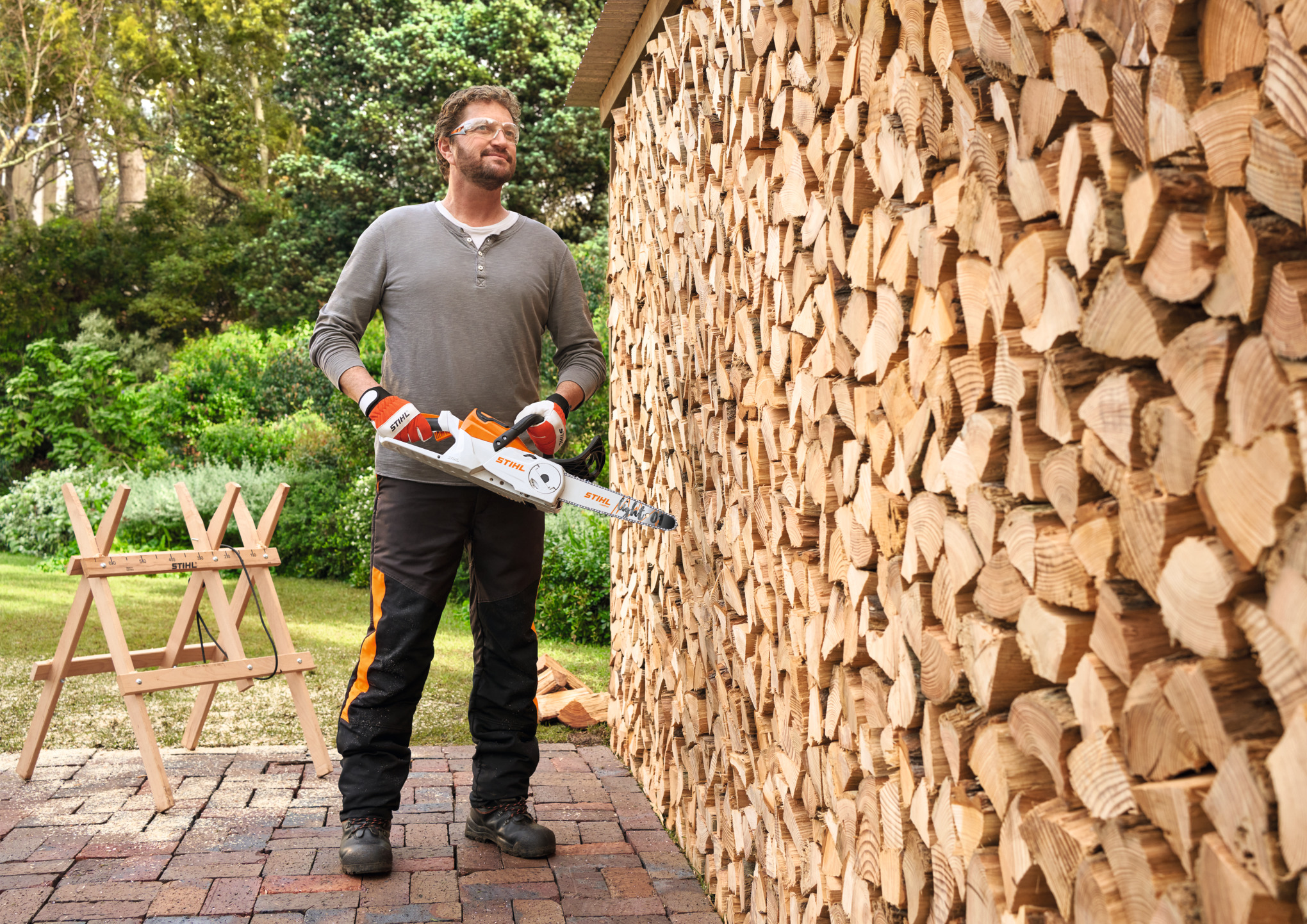 Un homme porte une tronçonneuse MSA 70 devant un mur de bois de chauffe