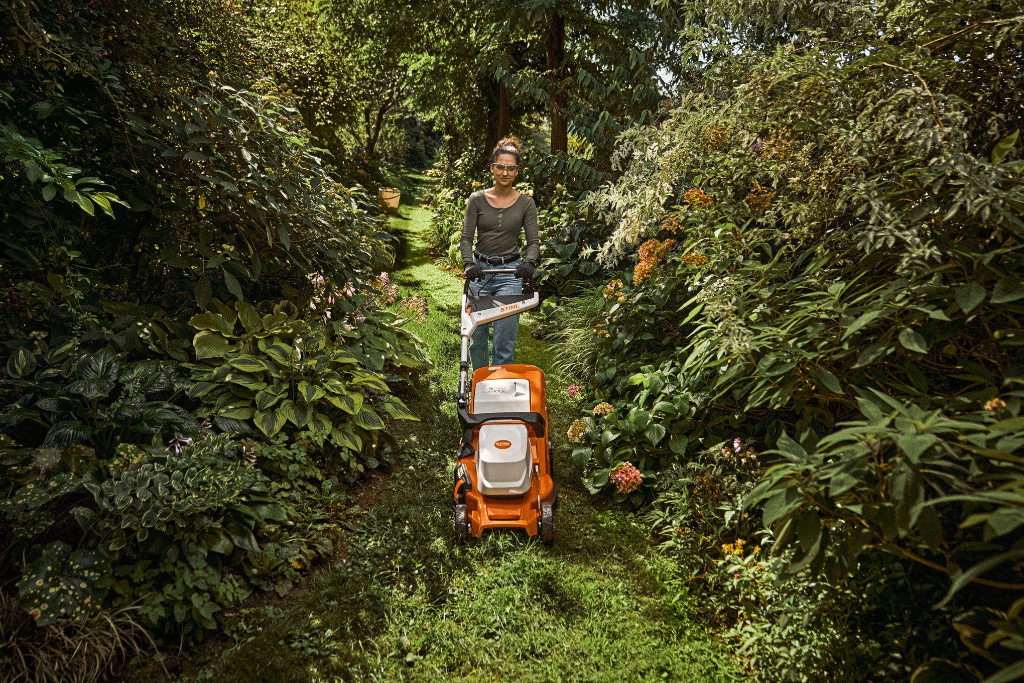 une femme tond la pelouse de son jardin entre 2 buissons avec une tondeuse à batterie STIHL