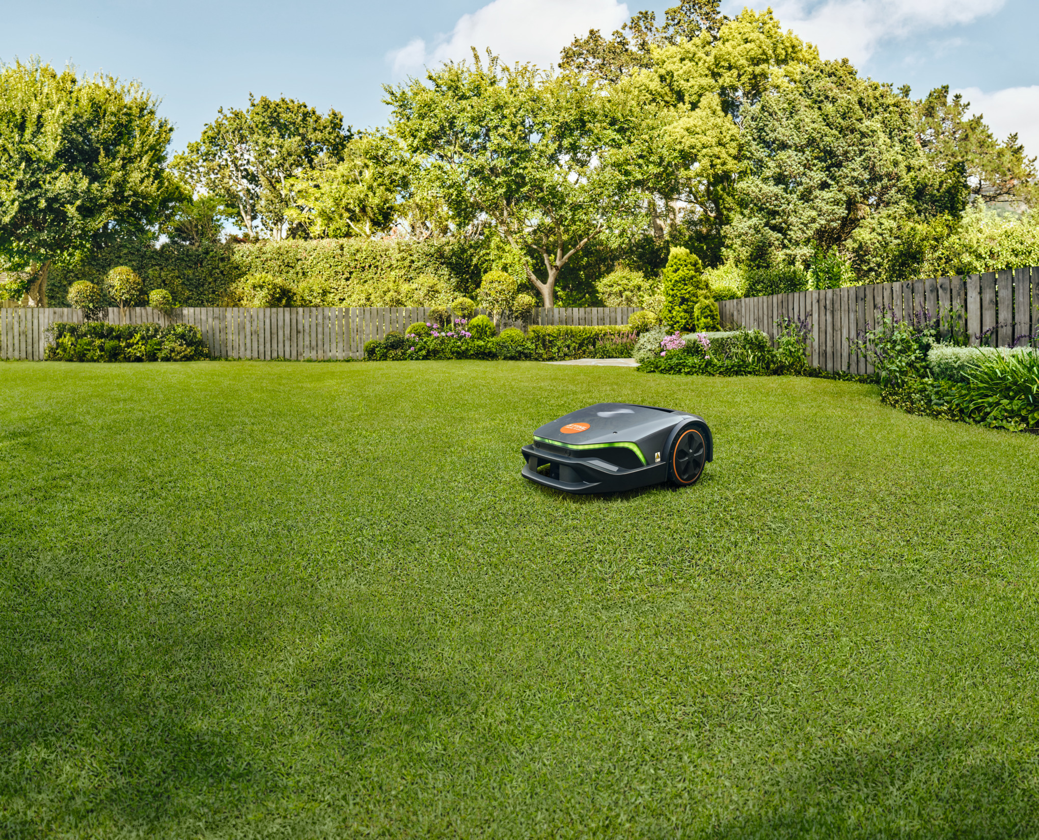 Un homme et un enfant jouent au football à l'arrière plan pendant qu'un robot tondeuse iMOW tond la pelouse