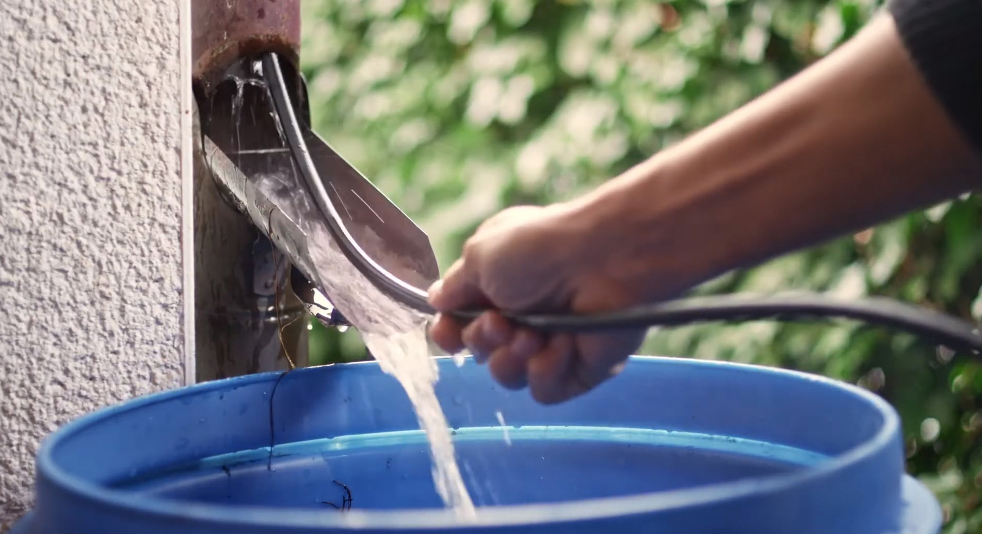 Une tige métallique flexible est poussée dans un tuyau de descente des eaux de pluie avec de l’eau s’écoulant dans un tonneau de récupération bleu