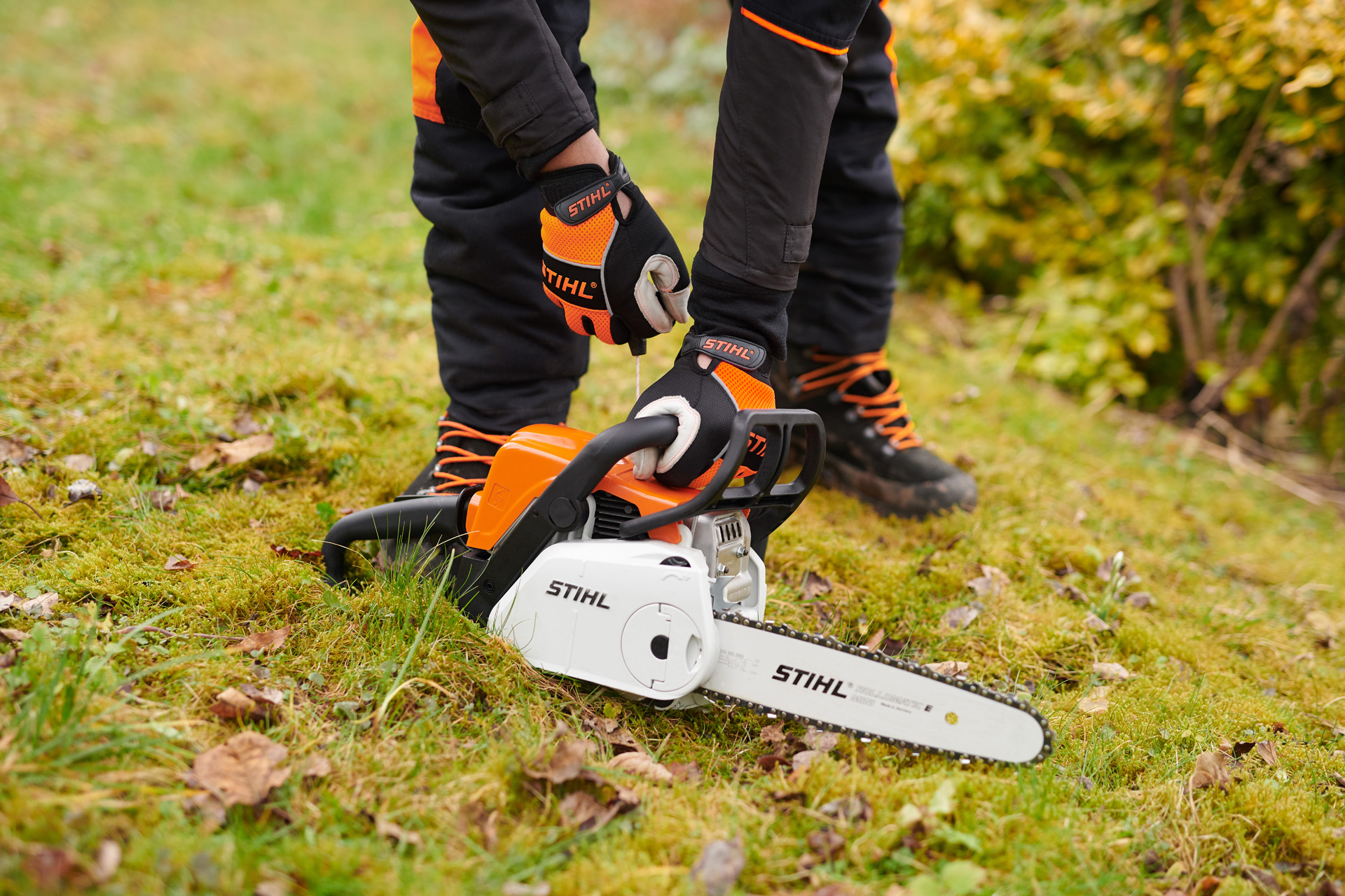 Une personne portant des gants de protection tient une tronçonneuse STIHL MS 180 C-BE au sol par la poignée.