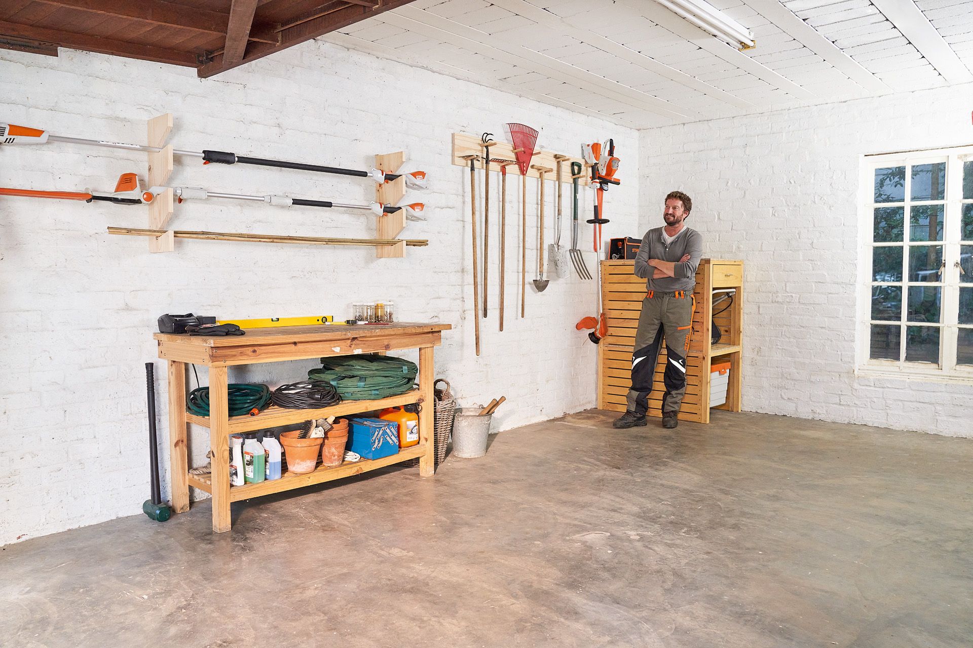 Un homme s’appuie les bras croisés sur un support de rangement pour outils de jardin fait maison dans un garage 