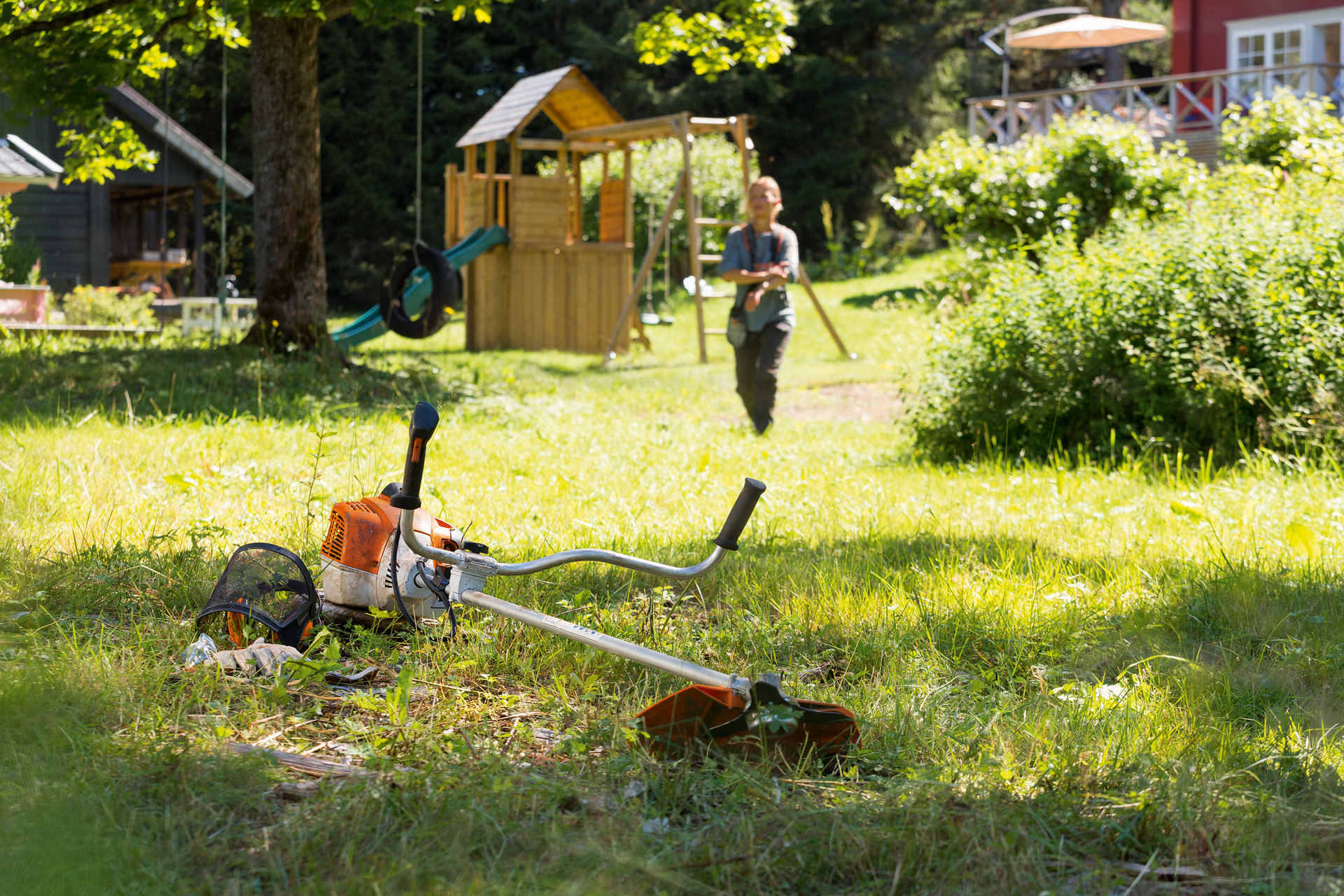 Débroussailleuse STIHL FS 240 au sol dans un jardin, avec des jeux pour enfant et un homme s’approchant de l’outil au loin
