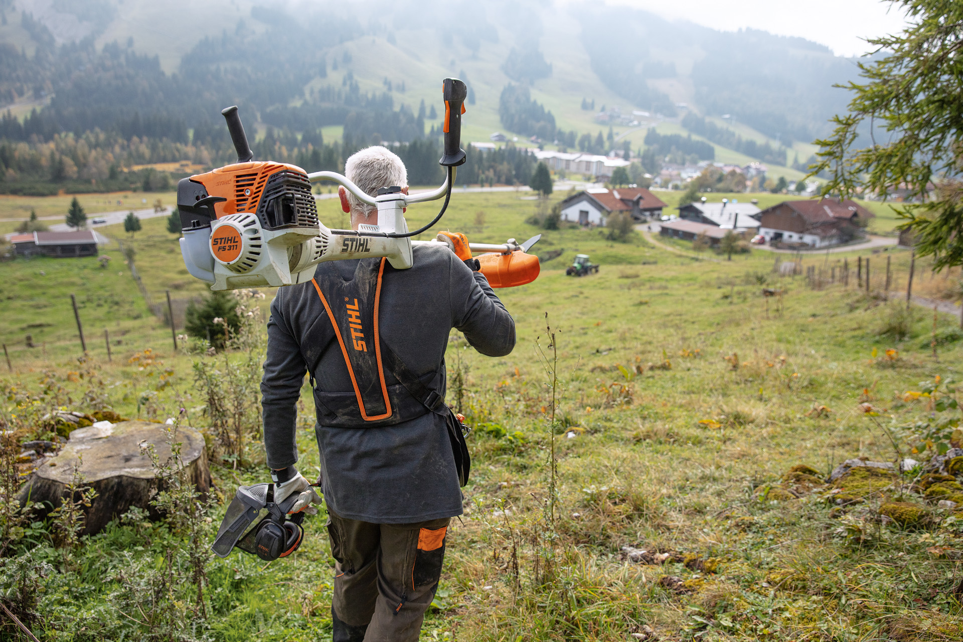 Un homme avec une scie de débroussailleuse STIHL FS 311 sur l’épaule, avec des collines vertes et des maisons en arrière-plan