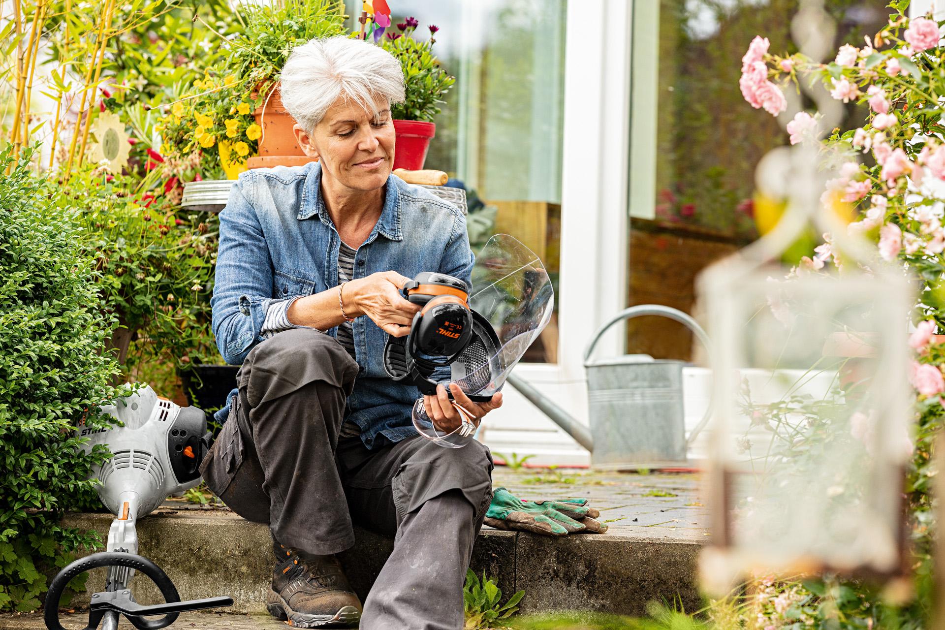 Une femme assise sur une marche dans un jardin, tenant une visière STIHL avec des protège-oreilles, avec un coupe-bordure sur le sol à proximité