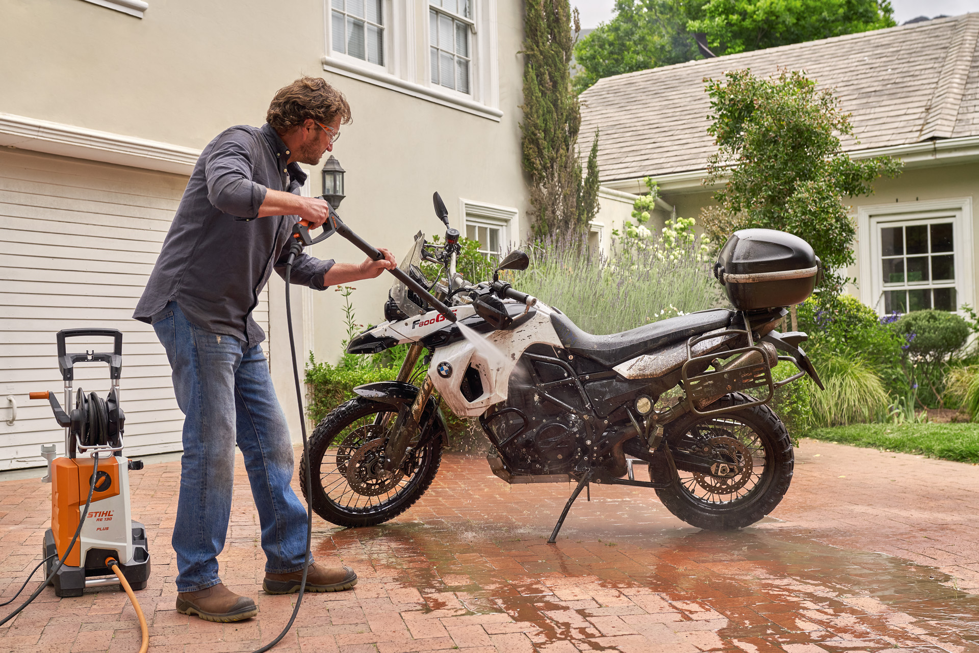 Homme utilisant un nettoyeur haute pression STIHL RE 130 Plus pour laver une moto blanche sur une allée de maison.