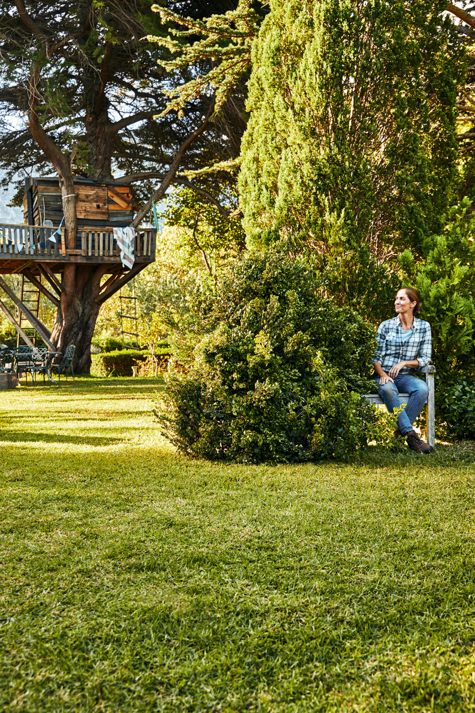 Une femme assise sur un banc dans un jardin entourée d’arbustes et de la pelouse, avec une cabane dans l’arbre en arrière-plan