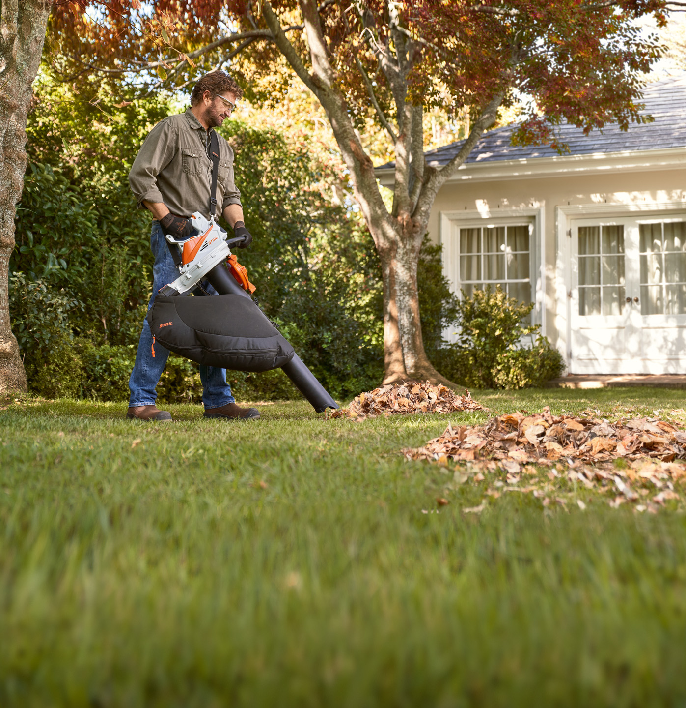 Un homme aspire les feuilles mortes d’un arbre dans son jardin avec l’aspiro-souffleur broyeur à batterie STIHL SHA 56