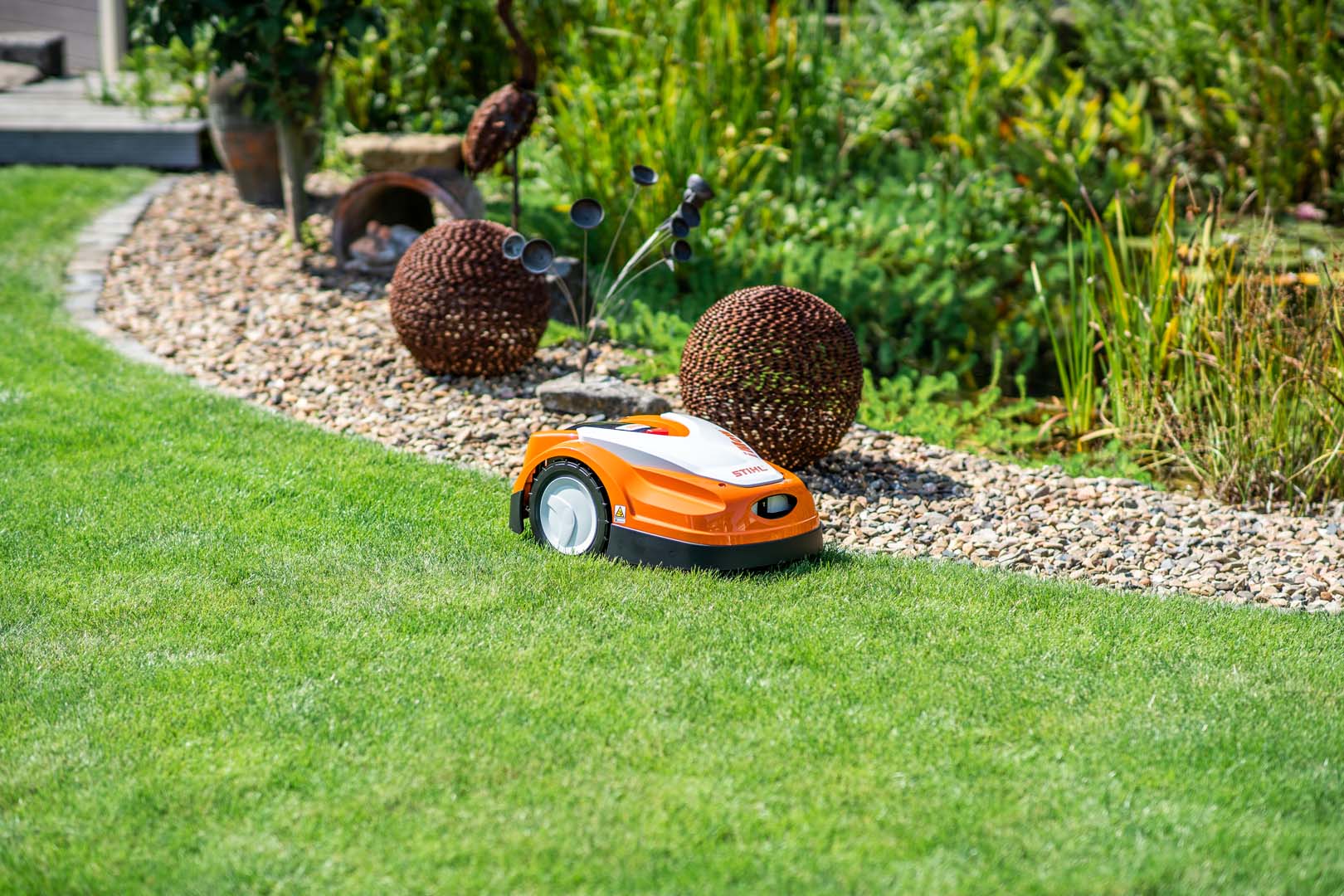 Un robot tondeuse STIHL iMOW® RMI 422 sur une pelouse à côté d’un lit de gravier et de sculptures de jardin