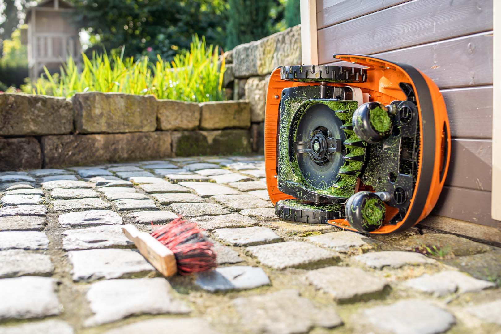 Un robot de tonte STIHL iMOW® tourné sur le côté, prêt à être nettoyé, avec une brosse posée juste à côté sur le sol