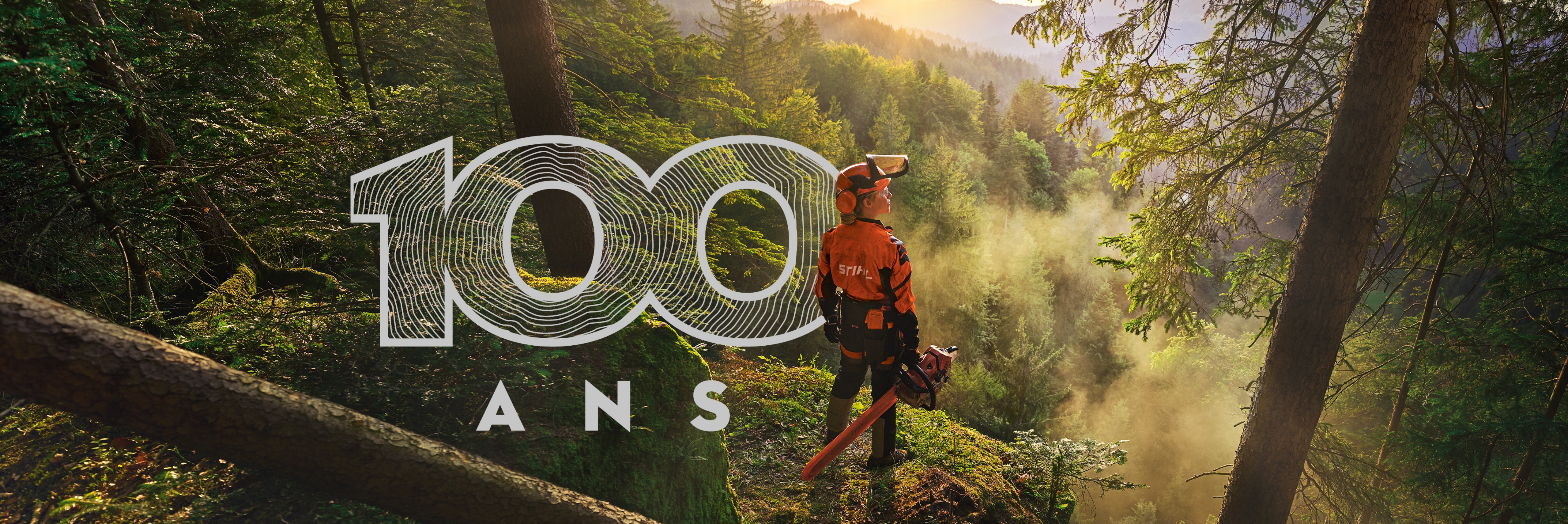 une femme bucheron tient à la main une tronçonneuse MS 500i en forêt face à la lumière et logo 100 ans