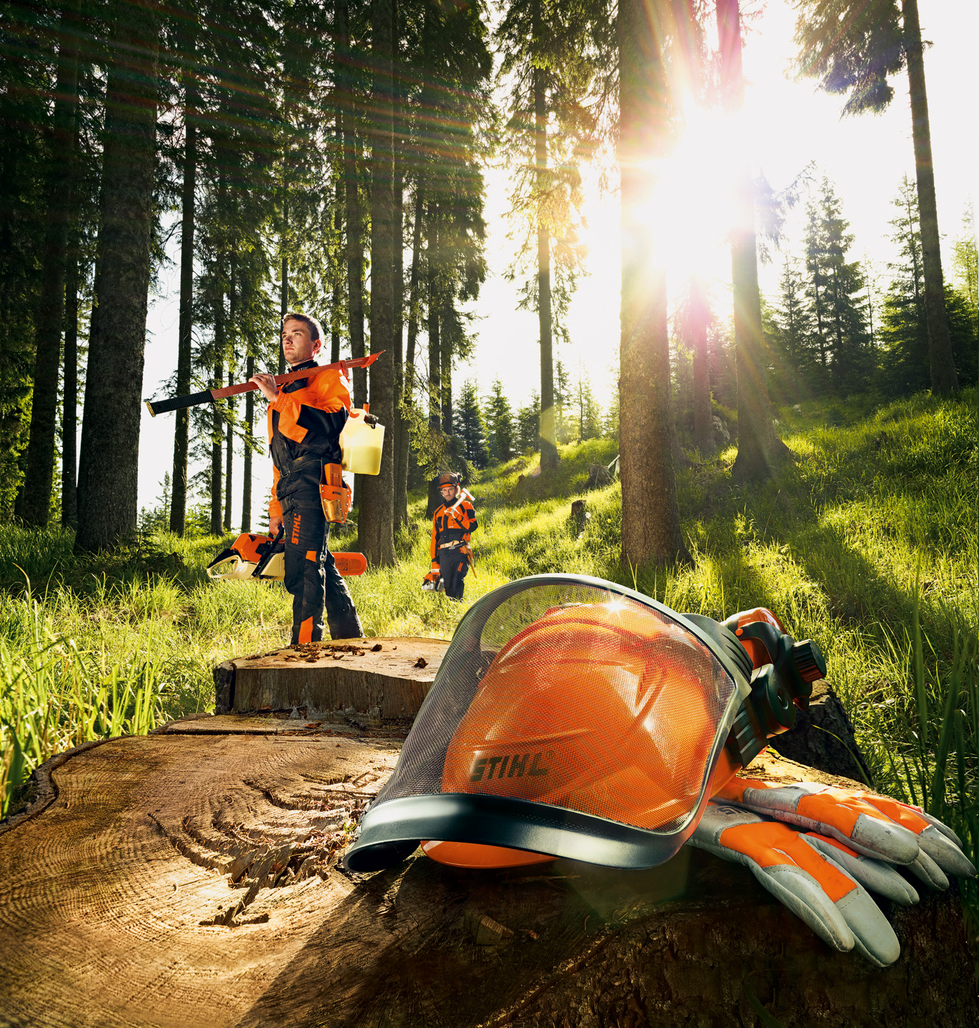 Deux bûcherons dans une forêt tenant des outils STIHL. Un casque au premier plan posé sur une souche d’arbre avec des gants de protection.