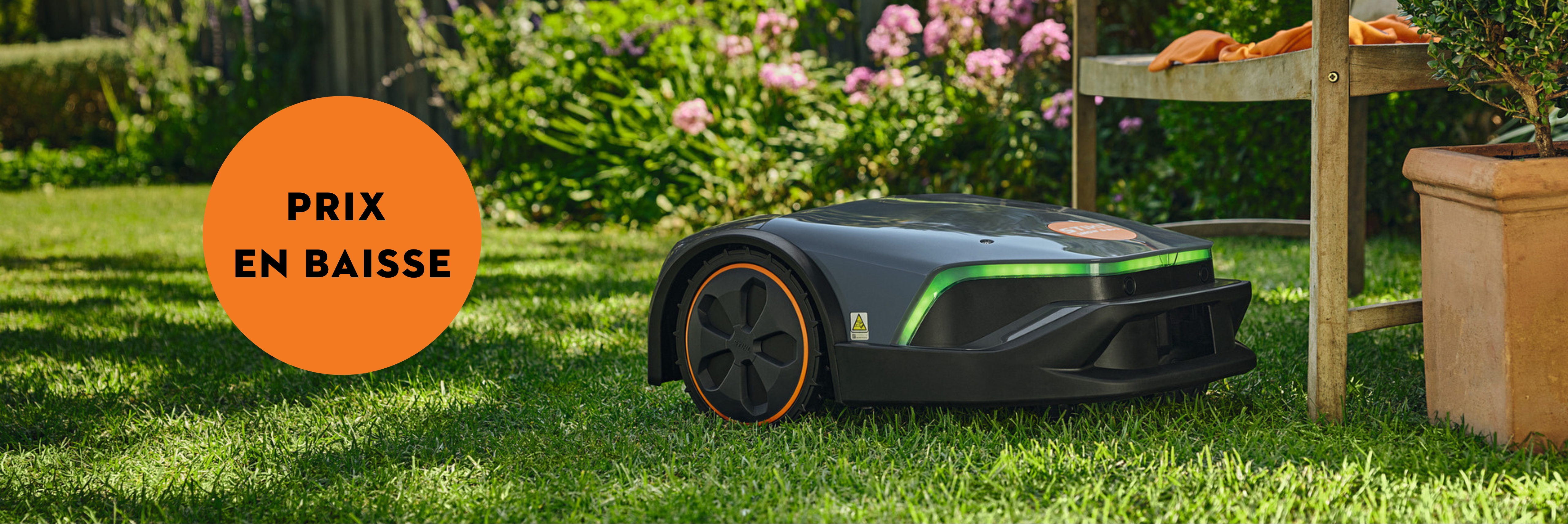 un robot tondeuse STIHL sur une pelouse devant un rosier, près d'un pot de fleur et d'un banc en bois