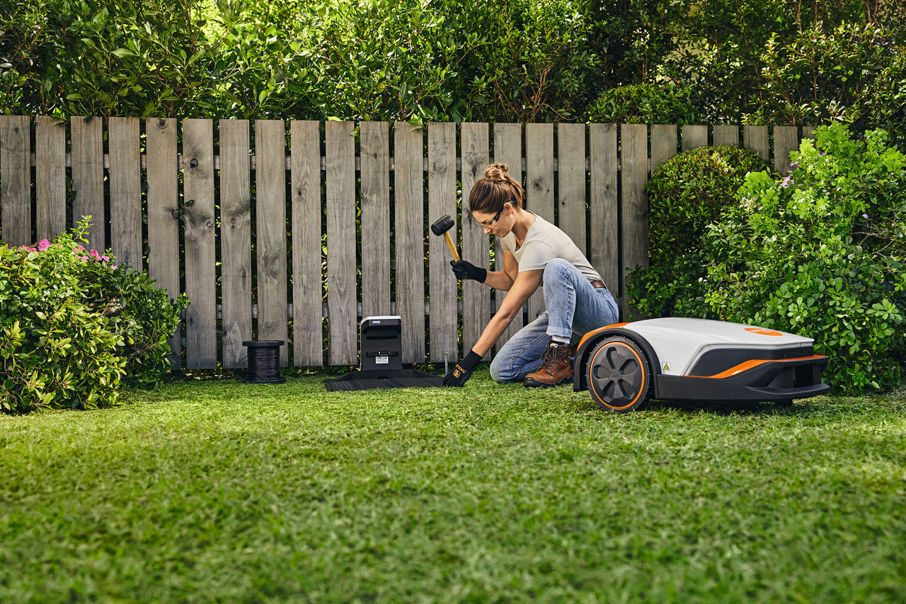 Femme dans un jardin, un marteau à la main, installant la station d’accueil iMOW®