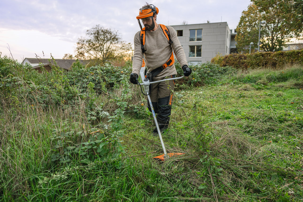Homme en équipement de protection STIHL tondant un espace vert envahi par les mauvaises herbes avec une débroussailleuse à batterie STIHL FSA 200