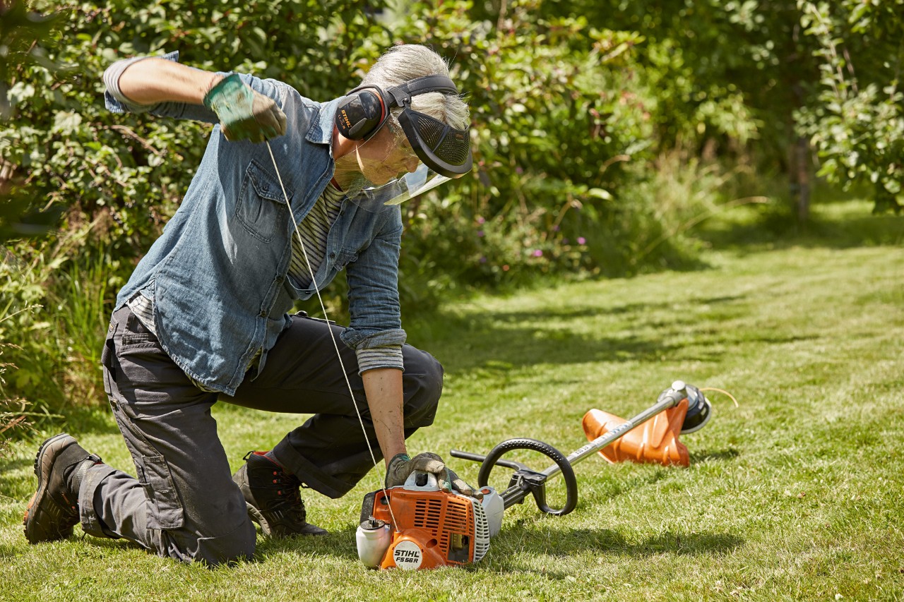 Femme portant des protections auditives et un masque en train de démarrer une débroussailleuse à essence STIHL FS 56 R sur un terrain vert
