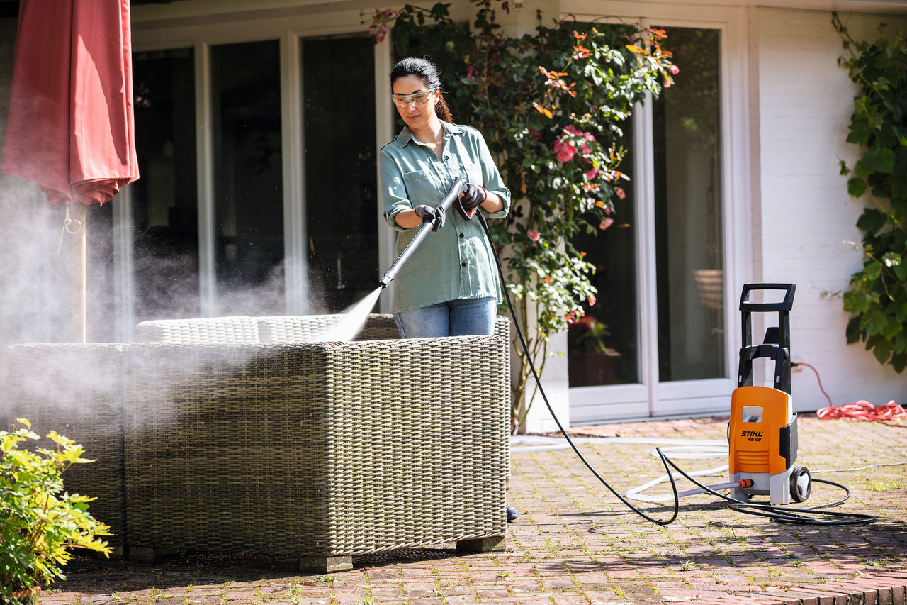 Femme portant des lunettes de protection nettoyant ses meubles de jardin avec le nettoyeur haute pression STIHL RE 88 sur sa terrasse.